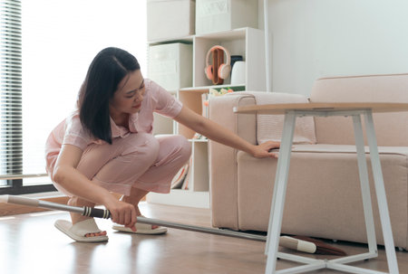 A woman is actively cleaning her living room, squatting beside a sofa to reach under furniture. The bright space features organized shelves and soft lighting, depicting a tidy home environment.の写真素材