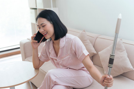 In a bright, modern living room, a young Asian woman engages in a cheerful phone conversation. She sits comfortably in pajamas, holding a cane, showcasing a relaxed morning atmosphere.の写真素材