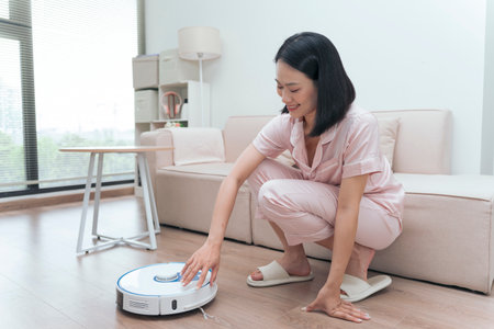A woman dressed in light loungewear is crouching down to operate a robotic vacuum cleaner in a bright, modern living room. Natural light streams through large windows, illuminating the space.の写真素材