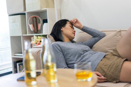 A woman is lying on a couch, holding her head with a pained expression. Nearby, glasses and bottles...
