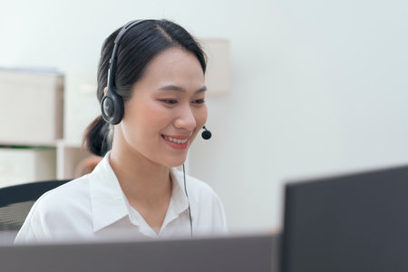 A focused Asian woman is engaged in her work at a desk, wearing a headset while smiling at her...