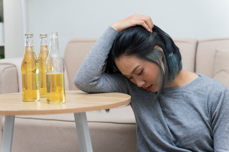 A young Asian woman in a gray sweater sits at a table, resting her head on her arm and looking down....