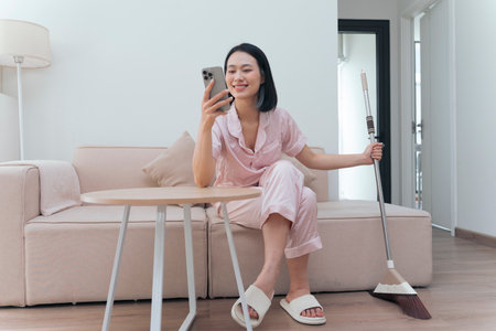 A woman sits comfortably in her stylish living room, dressed in light loungewear. She smiles while using her smartphone, holding a broom in her other hand, blending leisure and chores.の写真素材