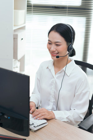 A young woman of Asian descent is smiling and engaging in a conversation while using a headset at her workstation. She sits at a desk, typing on a keyboard, and appears focused.の写真素材