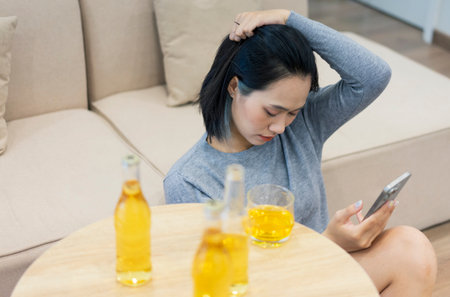 A young woman sits comfortably on a sofa while looking at her phone, sipping a drink from a glass. Several bottles of a beverage are on the table, creating a relaxed ambiance.の写真素材