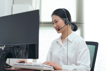 A focused woman is seated at her desk, wearing a headset and smiling while using a computer. Natural...