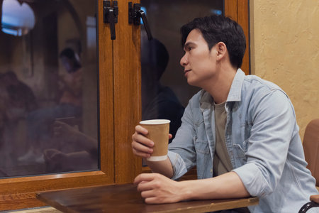 A man sits at a wooden table in a cozy cafe in Hanoi, Vietnam, holding a takeaway coffee cup. He gazes thoughtfully out the window, lost in contemplation during a quiet time.の写真素材