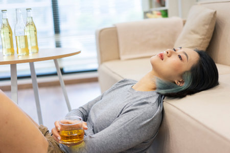 A young Asian woman sits on the floor against a sofa, holding a glass of drink. She looks relaxed,...