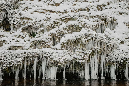 Wild river in the forest in winter with rocks and a stream of water and sandy cliffs with ice freezes and iciclesの写真素材