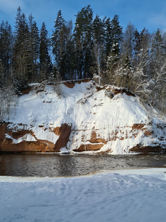 snow-covered cliff along which flows a small forest river. Green trees and blue sky above the cliff.Winter landscapeの写真素材