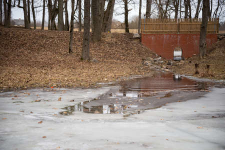 red brick bridge of the pipeline infrastructure with an arched culvert, through which a small forest river flows and flows into the park pond, which is frozen in early spring. Spring landscapeの写真素材