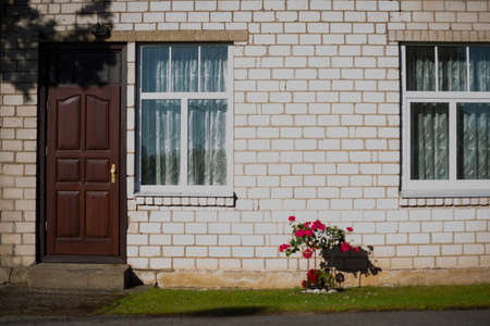 Red and white flowers in a flower pot with metal constructions on the white wall of a brick building with brown doors and wooden windows are lit in summer.の写真素材
