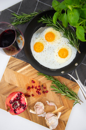 breakfast or brunch fried eggs in a black pan and red wine in a crystal glass and red pomegranate with garlic and basil. view from the top.の写真素材