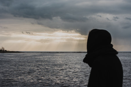 Close-up of a man dressed in black looking into the distance at the sea with a rocky pier with a white lighthouse on it.の写真素材