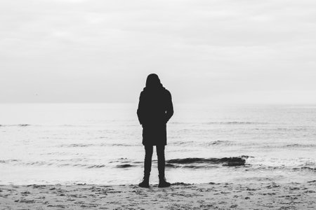depressed man in black clothes with a hood stands by the sea in the sand on the shore and looks into the distance.の写真素材