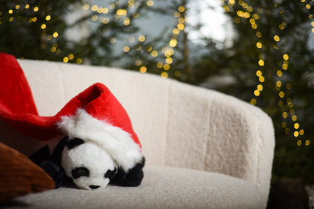 toy panda with a red gnome's Christmas hat is sitting on a white sofa near green fir trees in which the lights that create a bokeh effect are installed in the backgroundの写真素材