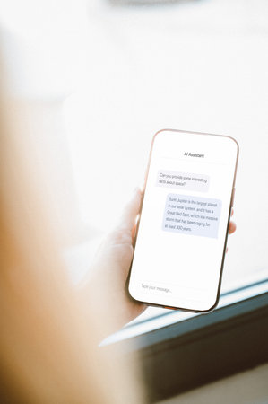 Close-up of a hand holding a smartphone displaying a modern AI assistant chat interface. Clean...