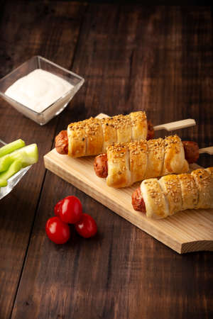 delicious snack of sausages wrapped in puff pastry with sesame seeds accompanied by cherry tomatoes, celery sticks and sauce. Served on wooden boardの写真素材