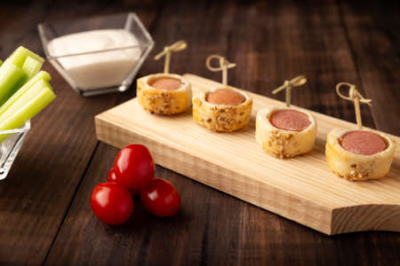 delicious snack of sliced sausages wrapped and in puff pastry with sesame seeds accompanied by cherry tomatoes, celery sticks and sauce. Served on wooden boardの写真素材