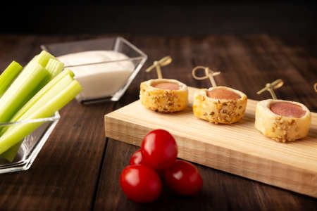 delicious snack of sliced sausages wrapped and in puff pastry with sesame seeds accompanied by cherry tomatoes, celery sticks and sauce. Served on wooden boardの写真素材