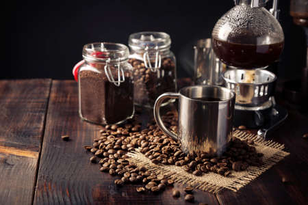Metallic cup and vacuum coffee maker also known as vac pot, siphon or syphon coffee maker and toasted coffee beans on rustic wooden tableの写真素材