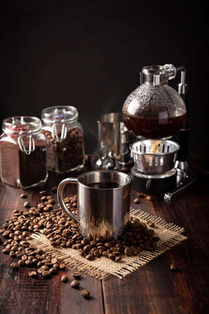 Metallic cup and vacuum coffee maker also known as vac pot, siphon or syphon coffee maker and toasted coffee beans on rustic wooden tableの写真素材