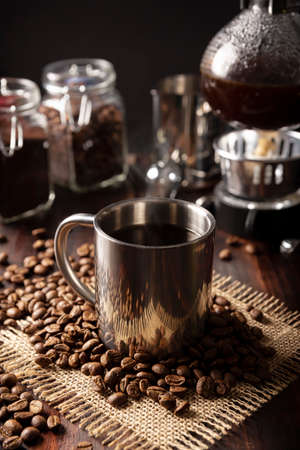 Metallic cup and vacuum coffee maker also known as vac pot, siphon or syphon coffee maker and toasted coffee beans on rustic wooden tableの写真素材