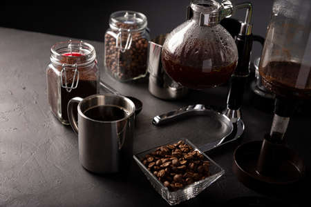 Vacuum coffee maker also known as vac pot, siphon or siphon coffee maker. Metallic cup and toasted coffee beans on rustic black stone table.の写真素材