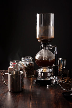 Vacuum coffee maker also known as vac pot, siphon or siphon coffee maker. Metallic cup and toasted coffee beans on rustic wooden tableの写真素材
