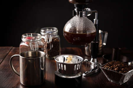Vacuum coffee maker also known as vac pot, siphon or siphon coffee maker. Metallic cup and toasted coffee beans on rustic wooden tableの写真素材