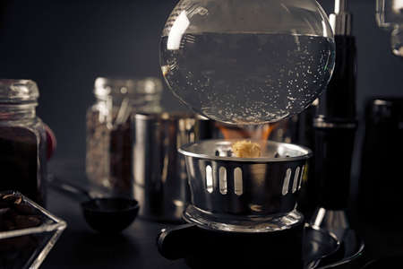 close up of lighter heating vacuum coffee maker also known as vac pot, siphon or syphon coffee maker on rustic black stone table.の写真素材