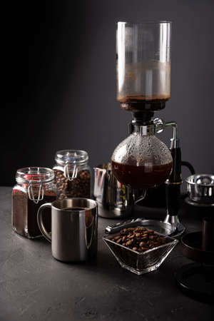 Vacuum coffee maker also known as vac pot, siphon or siphon coffee maker. Metallic cup and toasted coffee beans on rustic black stone tableの写真素材