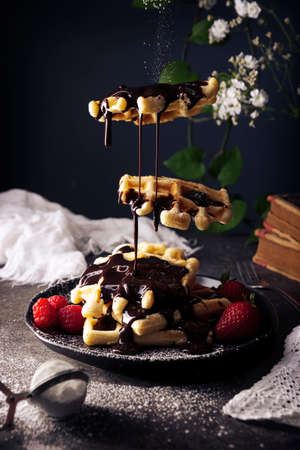 Creative image of homemade waffles levitating with liquid chocolate, fruits and powdered sugar on dark dish. Darkfood Levitation Photo.の写真素材