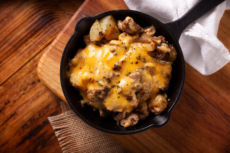 Homemade recipe for potatoes with steak and melted cheese, seasoned and served in a cast iron skillet, on a rustic wooden table. Table topview.の写真素材
