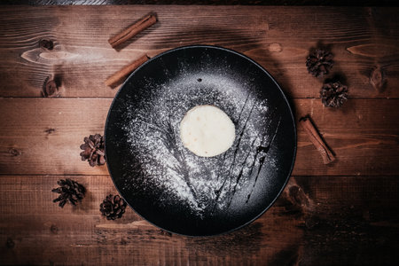 A round white mousse dessert, dusted with powdered sugar and drizzled with a dark sauce, is presented on a black plate, set against a wooden background. Top view, flat lay.の写真素材