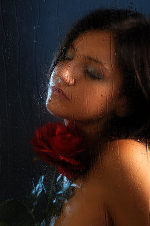 Beautiful young woman with a red rose behind a wet window.の写真素材