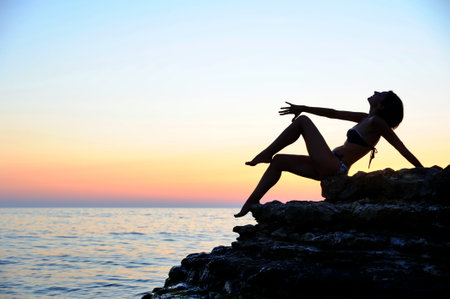 Silhouette of a beautiful young woman sitting on a rock by the sea at sunsetの写真素材