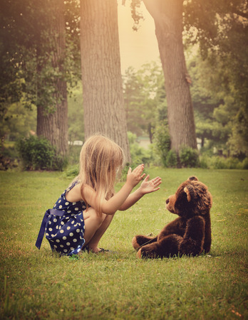 A little girl is clapping her hands and playing with a teddy bear outside at a park for a friendship or imagination concept. A little girl is clapping her hands and playing with a teddy bear outside at a park for a friendship or imagination concept.