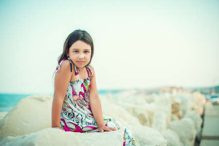 Beautiful little girl in dress sitting on the beach rocksの写真素材