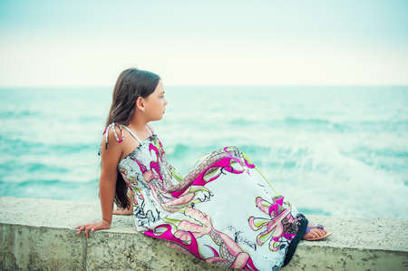 Beautiful little girl in dress sitting on the beach and looking at the horizonの写真素材