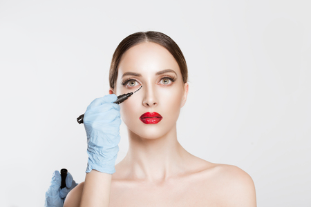Plastic surgery concept. Doctor Hands drawing marks on female face against white grey background. Facial Treatment. Portrait Of Beautiful Sexy Woman Black Surgical Lines around eyes skinの写真素材