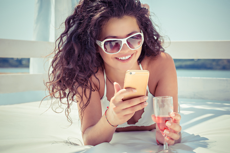 Closeup young beautiful happy, excited smiling woman texting, looking on mobile cell phone drinking champagne outdoors on sun-bed at sea, lake. Positive facial expression human emotion, feelings.の写真素材