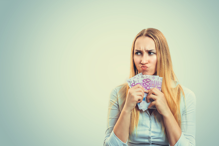 Blond woman in white shirt holding heap of money and frowning in greediness looking awayの写真素材