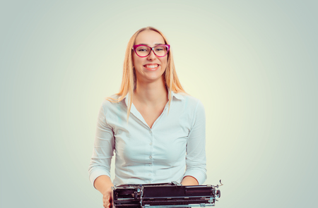 Happy young blond woman in glasses sitting with vintage typewriter smiling at cameraの写真素材