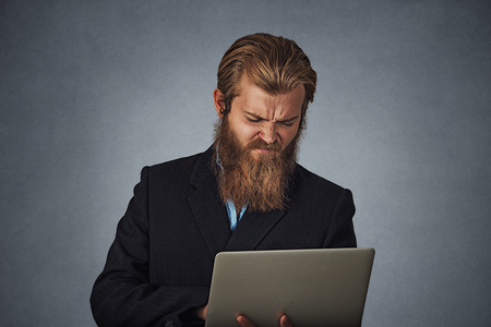 Bearded business man unhappy male holding laptop computer frowning isolated on gray grey studio wall Background. Negative face expression human emotion body language reaction attitude. Horizontal shotの写真素材