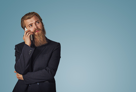 Bearded pensive hipster young businessman holding looking up talking speaking on phone thinking isolated on blue studio wall background. Serious face expression, human emotion, body language, reactionの写真素材