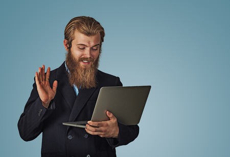 Hi there. Bearded hipster business man having a video call with a laptop greeting someone isolated on blue background with copy space. Multicultural model, mixed race asian russian caucasian model.の写真素材