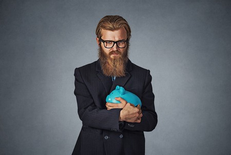 Young formal man in glasses extremely fascinated with money saving holding with greedy hand gesture the piggy bank. Bearded hipster businessman. Isolated on gray studio wall Background. Negative face.の写真素材