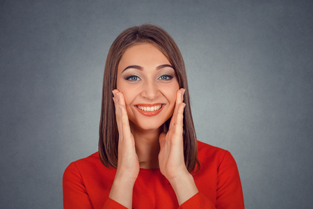 Closeup portrait of happy cute young woman in red dress looking excited surprised in full disbelief hands on cheeks isolated on gray grey studio wall background. Positive human emotion face expressionの写真素材