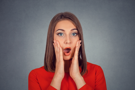 Shocked surprised stunned. Closeup portrait happy young woman in red looking excited in full disbelief hands on cheeks isolated on gray grey background. Positive human emotion face expression reactionの写真素材